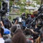 Eine große Gruppe von Journalisten und Kameraleuten hebt Videokameras und Mikrofone in die Luft, um bei einer Veranstaltung im Freien, umgeben von Grün und verschwommenen Menschen, Aufnahmen zu machen.