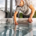 Ein Mann mit grauer Mütze und Polohemd prüft den Wasserstand und die Wasserqualität in einem Whirlpool im Freien, beugt sich vor und berührt das Wasser mit einer Hand.