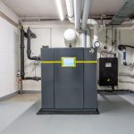 Eine moderne Industriemaschine mit Digitalanzeige steht in einem sauberen Technikraum mit sichtbaren Rohren und Schläuchen an Wänden und Decke.