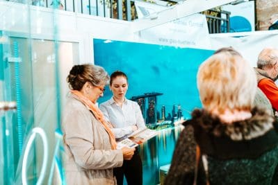 Eine junge Frau in einem weißen Hemd unterhält sich mit einer älteren Frau, die an einem Stand mit einer Stadtkulisse und mehreren anderen Ständen in der Nähe Broschüren in der Hand hält, über eine bevorstehende Pressemitteilung.