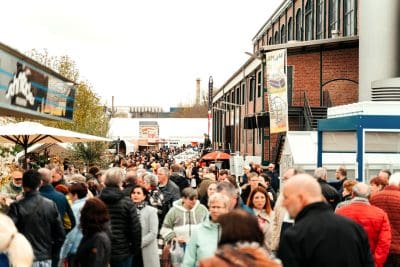 Eine große Menschenmenge schlendert durch einen Markt oder ein Festival im Freien, umgeben von Backsteingebäuden und Verkaufsständen unter bewölktem Himmel. Inmitten des geschäftigen Treibens sind bunte Fahnen und Regenschirme zu sehen, die eine lebhafte Atmosphäre schaffen, die einer Pressemitteilung würdig ist.