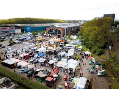 Luftaufnahme einer Freiluftveranstaltung mit vielen Zelten, Ständen und Fahrzeugen. Die Menschen flanieren zwischen den Auslagen auf einem großen Parkplatz in der Nähe von Industriegebäuden und Grünanlagen, im Hintergrund sind ein MediaMarkt und "Pressemitteilung"-Banner zu sehen.