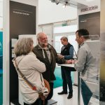 Ein Mann und eine Frau sprechen mit einem Aussteller an einem Messestand. Ein weiterer Mann steht in der Nähe, während im Hintergrund Messestände und Displays mit Branding zu sehen sind.