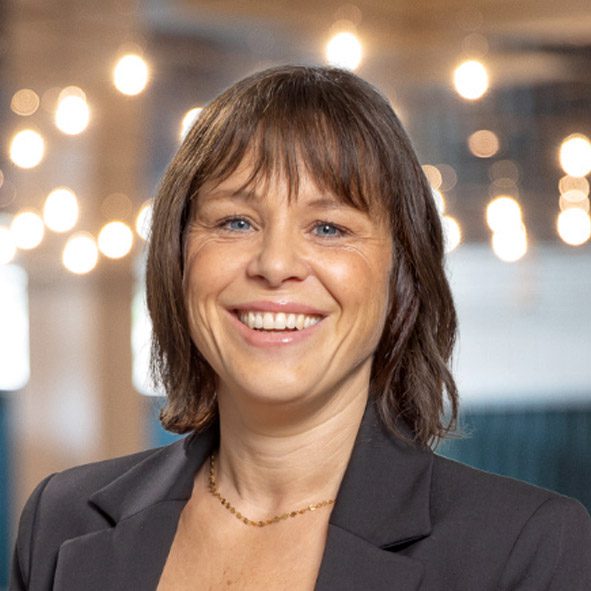 Eine Frau mit kurzen braunen Haaren und blauen Augen lächelt in die Kamera. Sie trägt einen schwarzen Blazer und eine dünne Halskette, mit verschwommenen Lichtern im Hintergrund.