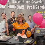Eine lächelnde Frau in einer Hornbach-Uniform steht an einem Stand und hält rosa Luftballons. Auf dem Tisch liegen Werbematerialien, orangefarbene Bleistifte und schwarze Tassen. Im Hintergrund ist ein großes Hornbach ProfiService-Banner zu sehen.
