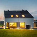 Modernes zweistöckiges Haus mit großen, in der Abenddämmerung warm leuchtenden Fenstern, schrägem Dach mit Oberlichtern und gepflegtem Rasen im Vordergrund.