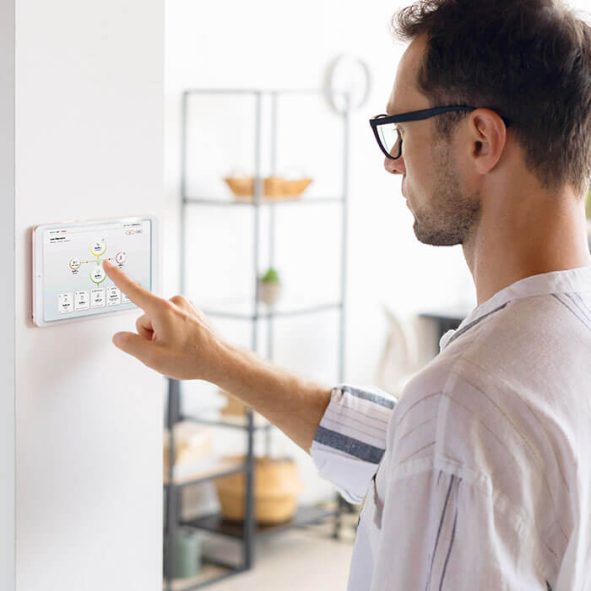Ein Mann mit Brille bedient ein an der Wand montiertes Touchscreen-Bedienfeld, wahrscheinlich für ein Smart-Home-System, in einem modernen, hellen Raum mit Regalen im Hintergrund.
