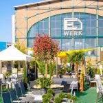 Sitzbereich im Freien mit Tischen, Stühlen und Topfpflanzen vor einem großen Backsteingebäude im Industriestil mit "E Werk"-Schriftzug und hohen Fenstern, unter blauem Himmel.