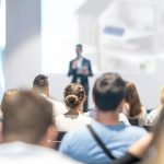 Das Publikum sitzt und hört einem Redner zu, der im vorderen Teil eines hellen Konferenzraums einen Vortrag hält, wobei der Redner und der Bildschirm im Hintergrund unscharf sind.