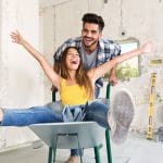Eine lächelnde Frau sitzt mit ausgestreckten Armen in einer Schubkarre, während ein Mann hinter ihr steht und die Karre schiebt. Sie wirken glücklich und verspielt in einem Raum, der gerade renoviert wird.