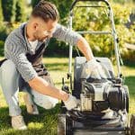 Ein Mann mit Schürze und Handschuhen kniet auf dem Rasen, während er in einem sonnigen Garten einen Rasenmäher wartet.