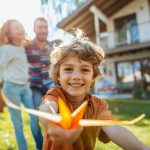 Lächelnder Junge, der ein Spielzeugflugzeug in einem sonnigen Hof hält, mit zwei Erwachsenen im Hintergrund in der Nähe eines Hauses, die alle einen hellen, fröhlichen Tag im Freien genießen.