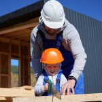 Ein Erwachsener und ein Kind, die beide Schutzhelme und blaue Overalls tragen, arbeiten gemeinsam mit einer Handsäge an einem Holzbrett auf einer Baustelle. Der Erwachsene führt das Kind, das lächelt, unter einem klaren blauen Himmel.
