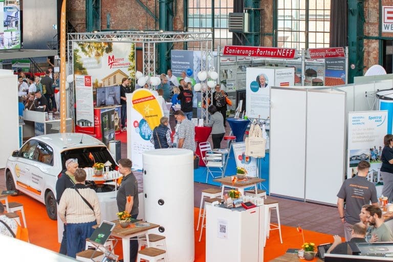 Eine belebte überdachte Messe mit Menschen, die in der Nähe von Ständen, Auslagen und einem weißen Auto herumlaufen und sich unterhalten. Bunte Banner, Informationsplakate und Tische mit Broschüren füllen die Szene. Die Umgebung wirkt lebendig und professionell.