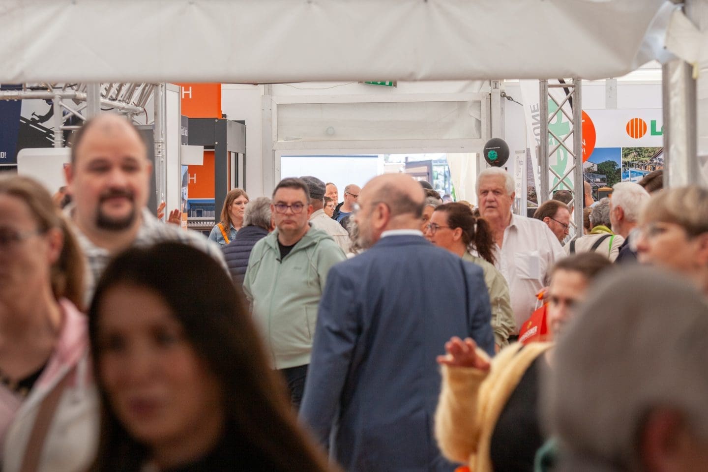 Eine Menschenmenge läuft durch eine belebte Ausstellung oder Messe mit Ständen und Auslagen im Hintergrund. Es herrscht eine lebhafte Atmosphäre mit Männern und Frauen unterschiedlichen Alters, die sich unterhalten.