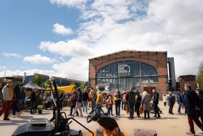 Vor einem großen Industriegebäude aus Backstein mit der Aufschrift E-WERK auf der Glasfassade versammeln sich unter einem teilweise bewölkten blauen Himmel eine Menschenmenge. Im Vordergrund sind Stände und Maschinen im Freien zu sehen.