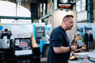 Ein Mann spricht an einem Stand auf einer Messe oder Ausstellung, um ihn herum liegen Ausstellungsmaterialien und Informationsschilder. Im Hintergrund interagieren mehrere Personen inmitten verschiedener Ausstellungsstände.