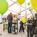 Drei Personen stehen um einen hohen Tisch herum und unterhalten sich in einem hellen Messestand. Im Hintergrund sind grüne Luftballons und Ausstellungsregale mit verschiedenen Artikeln zu sehen.