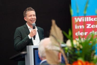 Ein Mann in einem grünen Anzug spricht lächelnd in ein Mikrofon an einem Podium. Im Vordergrund sind verschwommene Blumen zu sehen, und hinter ihm ist ein buntes Schild mit deutschem Text zu sehen.