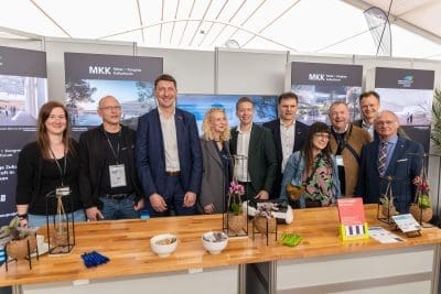 Eine Gruppe von neun Personen in Geschäftskleidung steht lächelnd an einem Messestand mit Werbedisplays hinter ihnen und einem Tisch mit Stiften, Süßigkeiten, Pflanzen und Broschüren vor ihnen.