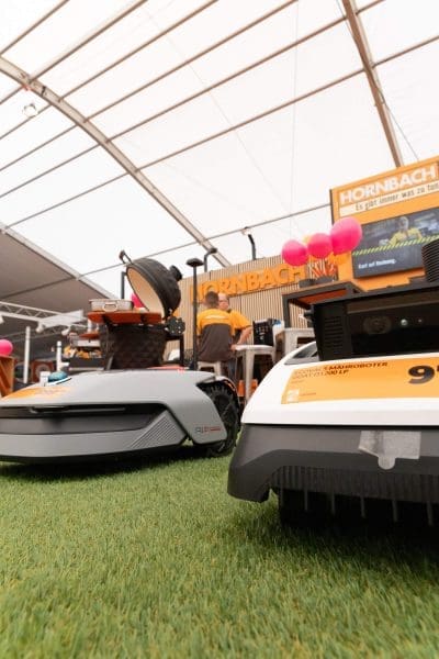 Zwei Roboter-Rasenmäher auf Kunstrasen bei einer Hornbach-Veranstaltung. Im Hintergrund sind eine Person und verschiedene Produkte in einem großen Zelt zu sehen.