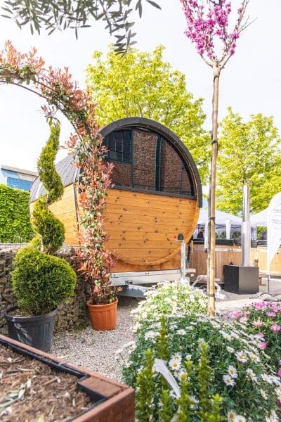 Ein rundes, hölzernes, fassförmiges Häuschen oder eine Sauna steht im Freien inmitten von Grün, Topfpflanzen und blühenden Blumen, mit einem dekorativen Torbogen und einem Baum in der Nähe.