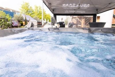 Nahaufnahme von sprudelndem Wasser in einem Whirlpool unter einem Vordach, mit Gartenmöbeln, Bäumen und Gebäuden im Hintergrund.