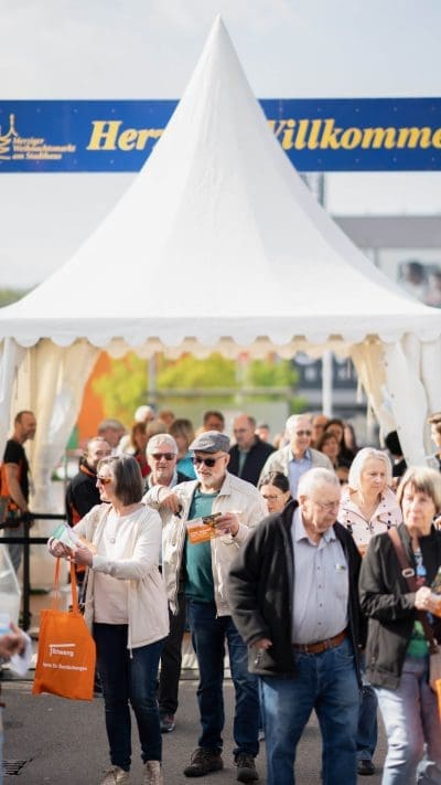 Eine große Gruppe von Menschen, zumeist ältere Erwachsene, steht und geht unter einem weißen Veranstaltungszelt mit einem blau-gelben Willkommen-Schild, das auf eine einladende Versammlung oder ein Fest im Freien hinweist.