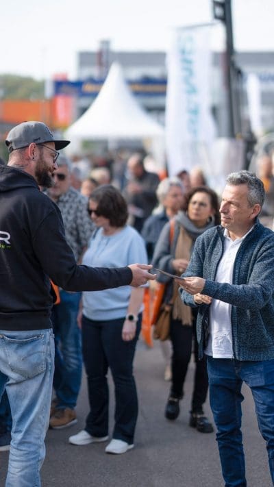 Ein Mann in einem schwarzen Kapuzenpulli überreicht einem anderen Mann in einem blauen Pullover ein Flugblatt, umgeben von einer Menschenmenge im Freien bei einer Veranstaltung oder einem Festival.