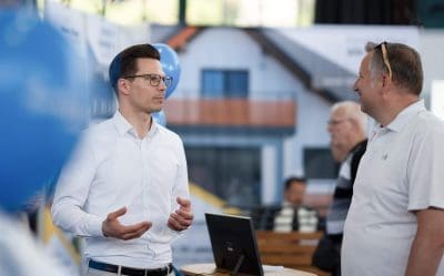 Zwei Männer stehen bei einer Veranstaltung in einer Halle und unterhalten sich. Ein Mann gestikuliert beim Sprechen, der andere hört zu und lächelt. Beide tragen Brillen und Freizeithemden. Im Hintergrund sind ein Laptop und ein verschwommenes Bild eines Hauses zu sehen.