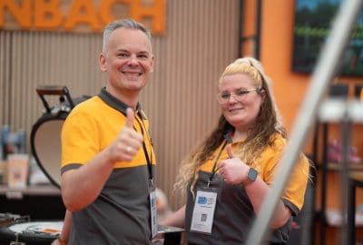 Zwei Personen in passenden orange-grauen Uniformen lächeln und zeigen den Daumen nach oben in die Kamera bei einer Indoor-Veranstaltung. Beide tragen Namensschilder und stehen vor einem unscharfen Hintergrund mit orangefarbener Beschilderung.