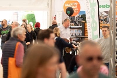 Eine geschäftige Veranstaltung in einer Halle, bei der verschiedene Menschen herumlaufen und sich unterhalten. Im Hintergrund sind Stände und Werbebanner zu sehen, und ein Mann in einem karierten Hemd steht hinter einem Tresen mit Broschüren.