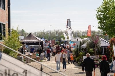 An einem sonnigen Tag spazieren die Menschen durch eine Freiluftmesse mit Verkaufszelten, Bannern, Luftballons und Pflanzenauslagen. Die Veranstaltung ist voller Besucher, und einige Schilder und Fahnen sind über den Zelten zu sehen.