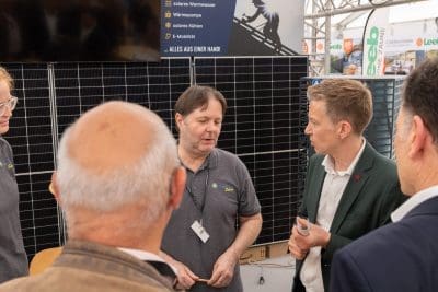 Ein Mann in einem grauen Polohemd erklärt einer Gruppe von Menschen an einem Solarenergiestand etwas, im Hintergrund sind Solarzellen und Informationsschilder zu sehen.