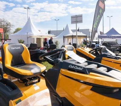 Mehrere gelbe Cub Cadet-Aufsitzrasenmäher werden auf einer Messe im Freien ausgestellt, mit weißen Zelten, Fahnen und Menschen, die im sonnigen Hintergrund interagieren.