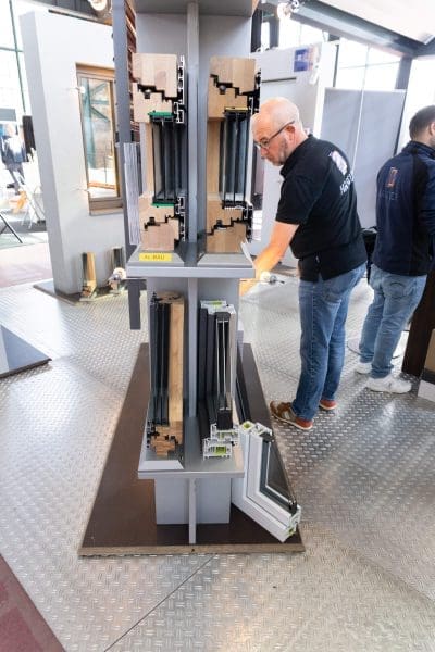 Ein Mann begutachtet Modelle von Fensterrahmen, die in einem Raum ausgestellt sind und verschiedene Schichten und Materialien zeigen, die im modernen Fensterbau verwendet werden. Eine weitere Person steht in der Nähe.
