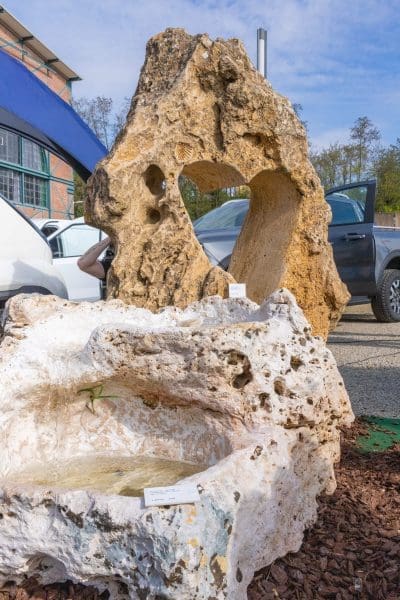 Große dekorative Felsen im Freien; der Felsen im Hintergrund hat ein herzförmiges Loch, während der Felsen im Vordergrund weiß ist und eine flache Schale hat. Im Hintergrund sind Autos und ein Gebäude zu sehen.