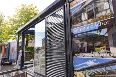 Eine moderne Glaskonstruktion mit horizontalen Jalousien wird im Freien ausgestellt. Werbeschilder in deutscher Sprache werben für Terrassendächer mit integrierten Geländern, in denen sich Bäume und Gebäude in der Nähe in den Glasscheiben spiegeln.