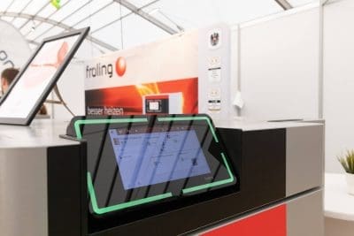Nahaufnahme eines Touchscreen-Bedienfelds mit grünem Rand auf einer Heizungsanlage auf einer Messe. Der Hintergrund zeigt einen Stand mit Froling-Branding und Informationspostern.