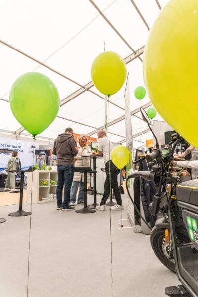 Menschen stehen um Stehtische herum und unterhalten sich bei einer Indoor-Veranstaltung oder -Ausstellung. Im Vordergrund schweben grüne und gelbe Luftballons, und im Hintergrund sind Displays und ein Motorroller zu sehen.
