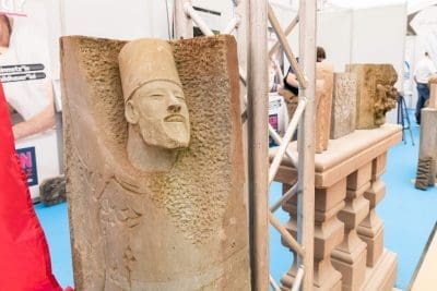 Eine Steinskulptur mit einem geschnitzten menschlichen Gesicht und einem Hut steht aufrecht neben anderen geschnitzten Steinfiguren in einer Innenausstellung mit blauem Boden und Metallstützen.
