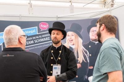 Drei Männer unterhalten sich an einem Veranstaltungsstand. Ein Mann trägt eine traditionelle schwarze Uniform und einen Hut, während die beiden anderen legere Kleidung tragen. Im Hintergrund ist ein Werbebanner mit Menschen in ähnlichen Uniformen zu sehen.