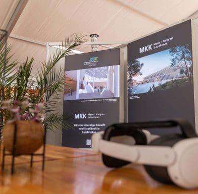 Ein Messestand mit zwei großen Plakaten, die architektonische Renderings für das MKK Messe | Kongress | Kulturforum in Saarbrücken zeigen. Ein VR-Headset und eine kleine Pflanze stehen auf dem Holztisch im Vordergrund.