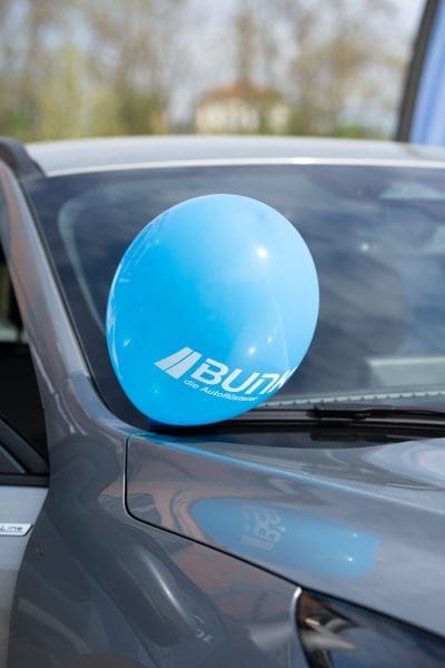 Ein blauer Luftballon mit weißem Text ist an den Seitenspiegel eines grauen Autos gebunden, wobei Bäume und Himmel im Hintergrund verschwimmen.