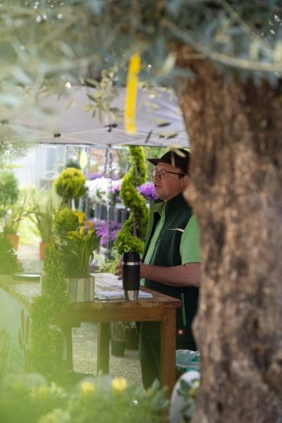 Eine Person mit Brille und grüner Weste steht hinter einem Holztisch an einem Marktstand im Freien, umgeben von Pflanzen und Blumen, die teilweise von einem Baum im Vordergrund verdeckt werden.