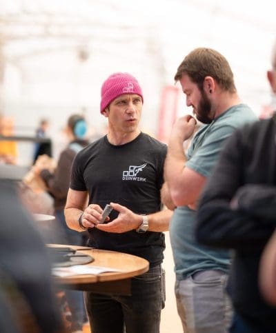 Zwei Männer unterhalten sich an einem runden Tisch. Der eine trägt eine rosa Mütze und ein schwarzes T-Shirt mit einem Logo und hält einen Gegenstand in der Hand. Der andere, in einem blauen Hemd, hört mit der Hand am Kinn zu. Der Hintergrund ist unscharf.