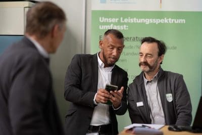 Zwei Männer in Geschäftskleidung stehen zusammen, einer zeigt dem anderen etwas auf seinem Handy, während eine dritte Person in der Nähe steht. Sie befinden sich in einem Innenraum, vor einem grünen Hinweisschild mit deutscher Aufschrift.