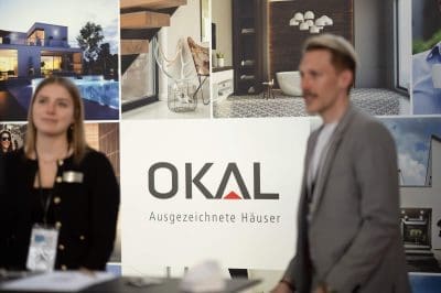Zwei Personen stehen vor einem Display mit dem OKAL-Logo und der deutschen Aufschrift Ausgezeichnete Häuser, auf dem im Hintergrund Fotos von modernen Häusern und Inneneinrichtungen zu sehen sind.
