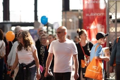 Eine Gruppe von Menschen geht auf einer Veranstaltung oder einem Jahrmarkt nach drinnen. Ein Mann in einem weißen T-Shirt und mit Sonnenbrille steht in der Mitte. Andere tragen orangefarbene Taschen und Luftballons, im Hintergrund ist ein rotes Marktkauf-Banner zu sehen.