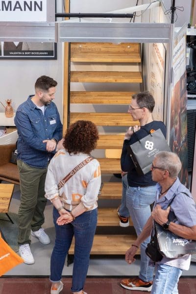 Vier Personen stehen am Fuße einer Holztreppe in einem Innenraum und unterhalten sich. Drei sind einander zugewandt, während eine Person mit einer Tasche vorbeigeht. Die Szene scheint auf einer Veranstaltung oder Ausstellung zu spielen.